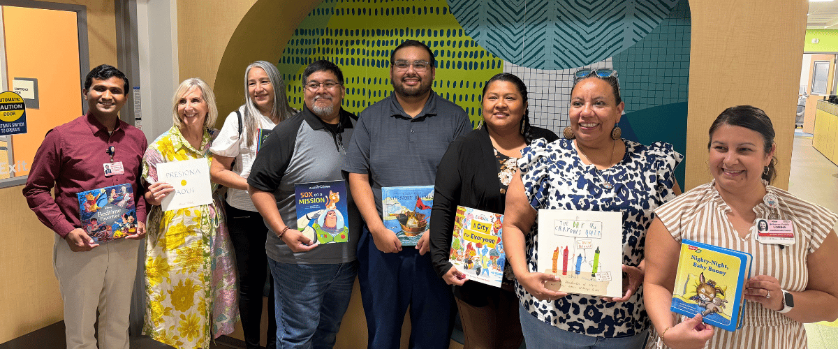 A diverse group of people stand in a hallway holding children's books, including "Press Start!," "Sox on Mission," "Everyone is Everyone," "My No, My No, My No," and "Nighty-Night, Baby Bunny."