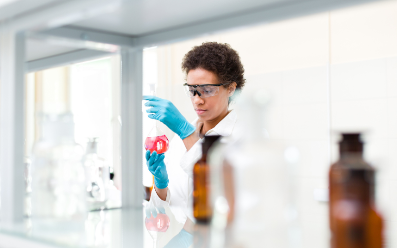 A person wearing protective goggles and blue gloves holds a flask with red liquid in a laboratory setting.