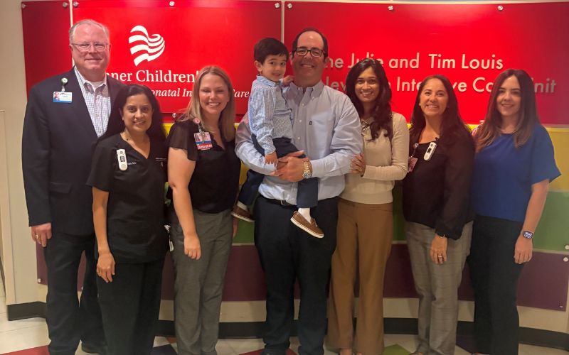 A group of medical professionals stand with a family in front of a red wall with text that reads 