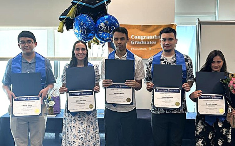 Five graduates stand in a line, each holding a diploma and wearing a blue sash over their shoulders. In the background, balloons and a banner that reads 