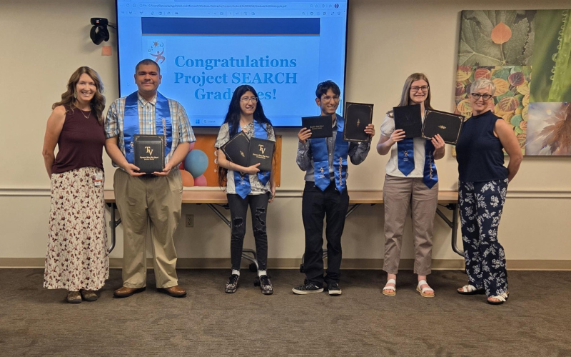 Four graduates, two women and two men, stand holding diplomas and wearing blue sashes, with two women standing on either end of the group.