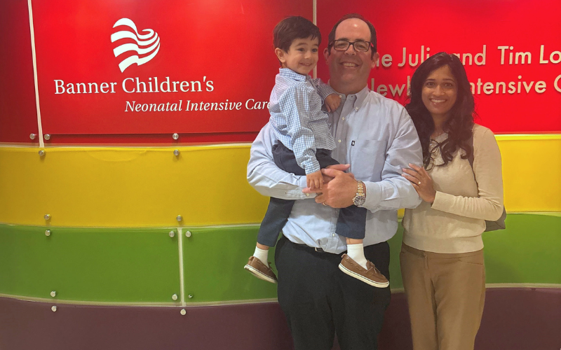 A family smiles in front of a red sign that reads 