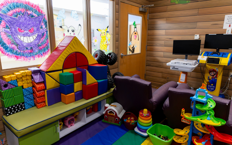 A colorful playroom with large soft blocks, toys, and screens on stands. Drawings of cartoon characters are on the window.