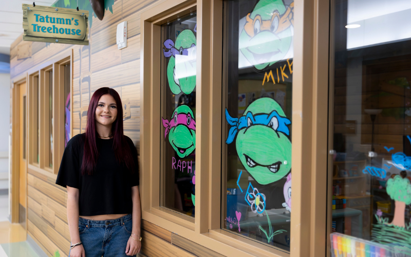 Tatumn Carr stands next to a window decorated with drawings of the Teenage Mutant Ninja Turtles, with a sign above reading 
