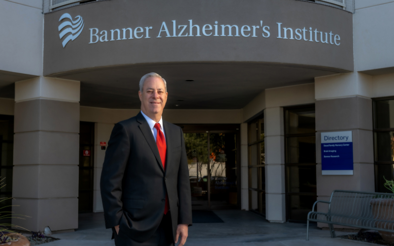 A man in a dark suit and red tie stands outside the Banner Alzheimer’s Institute entrance, with the institute name on the building above him and a blue directory sign to the right.