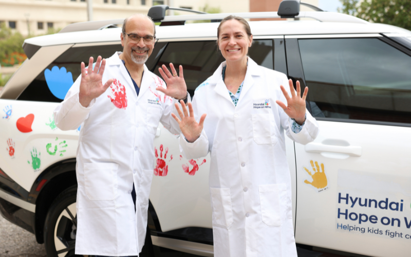Two healthcare workers in white lab coats wave at the camera beside a white van covered with colorful handprints and the words “Hyundai Hope on Wheels.”