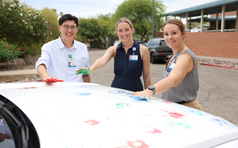 Three smiling staff members stand outdoors beside a car with a colorful painted hood, one in a white shirt and two in dark tops, with a campus building and parked vehicles in the background.