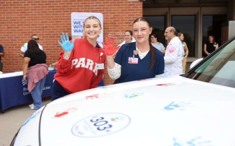 Two people in event badges stand behind a white car, smiling and waving with raised hands, in front of a brick building with other attendees in the background.