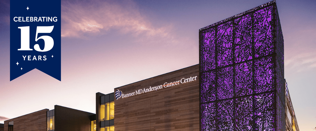 Modern university building at dusk with warm-lit windows and a tall facade covered in purple LED panels, framed by a landscaped walkway and trees.