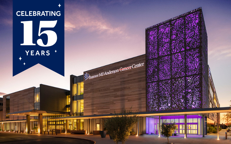 Modern university building at dusk with warm-lit windows and a tall facade covered in purple LED panels, framed by a landscaped walkway and trees.