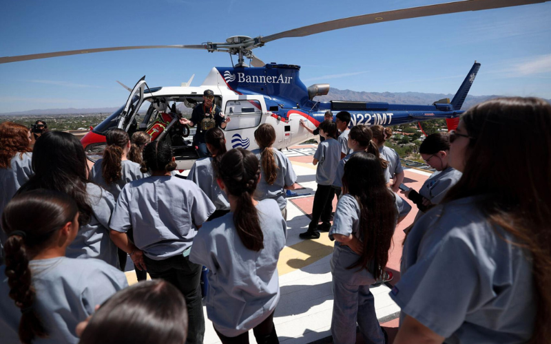 A crowd of students in light-blue shirts watches as military personnel stand on and around a gray aircraft on a runway, with a banner reading “BANNER” on the nose against a clear blue sky.