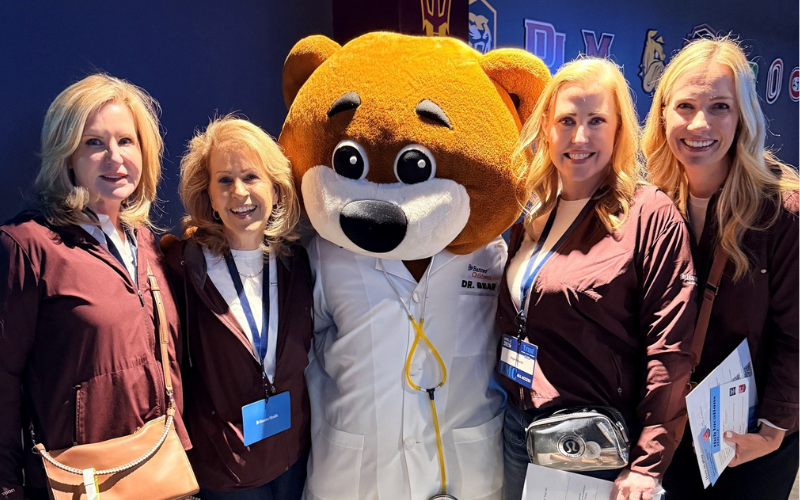 A mascot bear in a lab coat with a stethoscope poses with four women wearing maroon jackets.