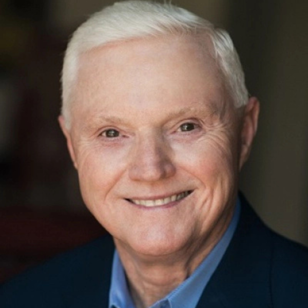A headshot of a smiling man with white hair, wearing a dark blue suit jacket over a light blue collared shirt.