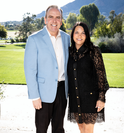A man in a light blue blazer and a woman in a black lace dress stand together outdoors with a golf course and mountains in the background.