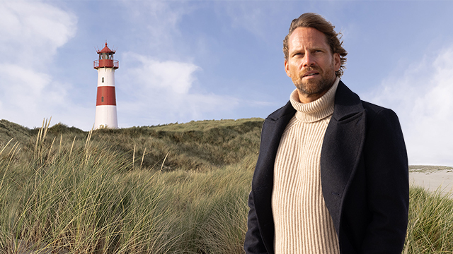 Mann steht an der Nordseeküste, im Hintergrund ein rot-weißer Leuchtturm
