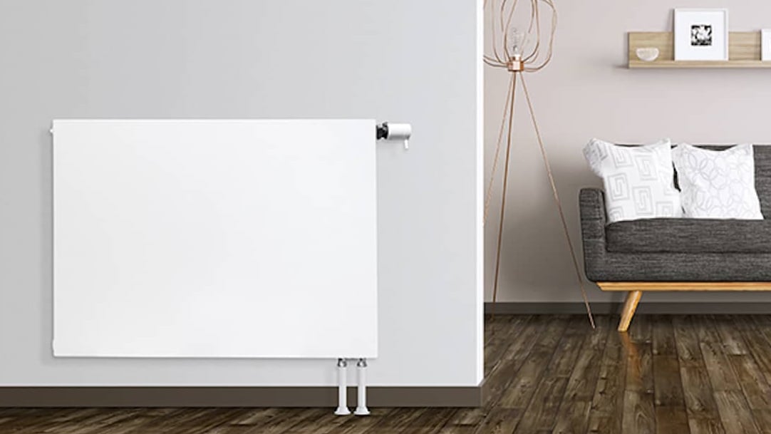 Ein EuroPlan-Flachheizkörper an einer hellgrauen Wand, rechts ein Wohnzimmer mit einer Couch.