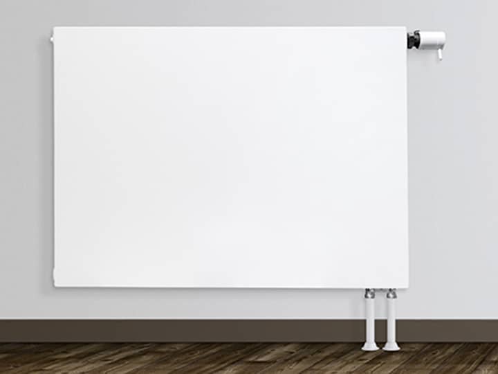 Ein weißer Flachheizkörper an einer hellgrauen Wand, rechts davon das Wohnzimmer mit Couch, Tisch und Wandregal.