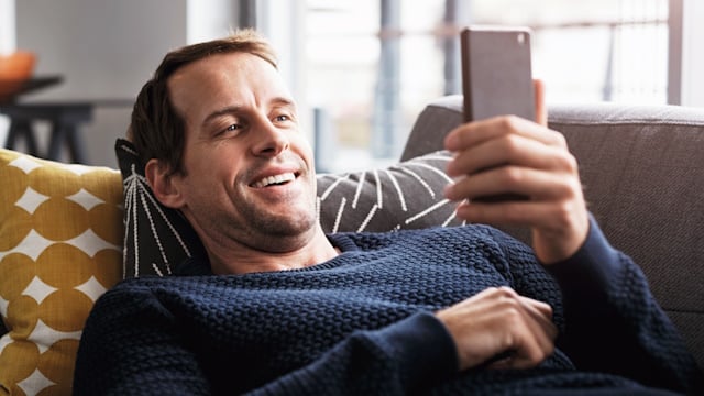 Mann liegt auf der Couch, schaut auf sein Handy und informiert sich online über Förderungen rund um Wärme
