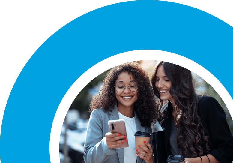 Two woman smiling as one is holding her phone and coffee and her friend looks over to her screen