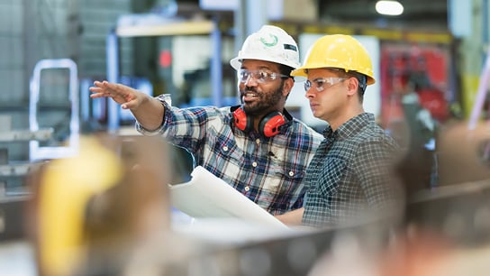 Two men equipped with hard hats and safety glasses are attentively inspecting an item, emphasizing workplace safety protocols.