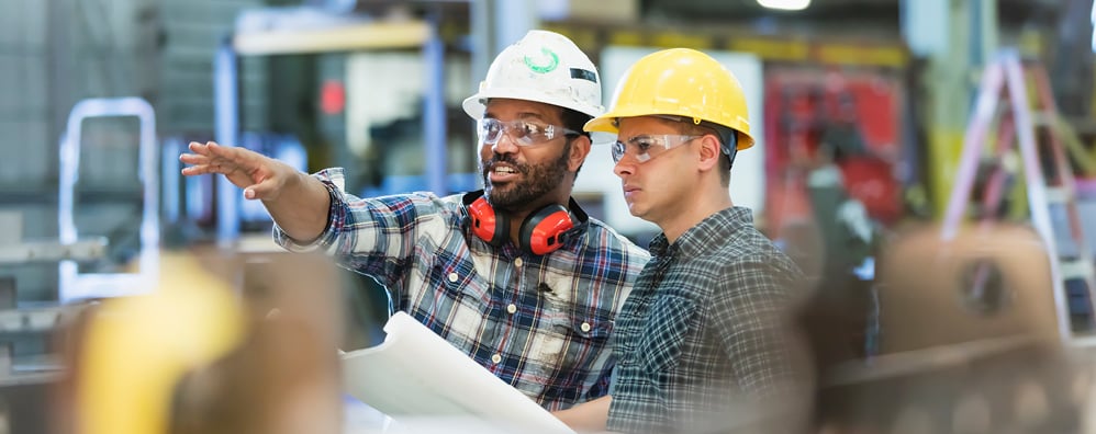 Two men equipped with hard hats and safety glasses are attentively inspecting an item, emphasizing workplace safety protocols.