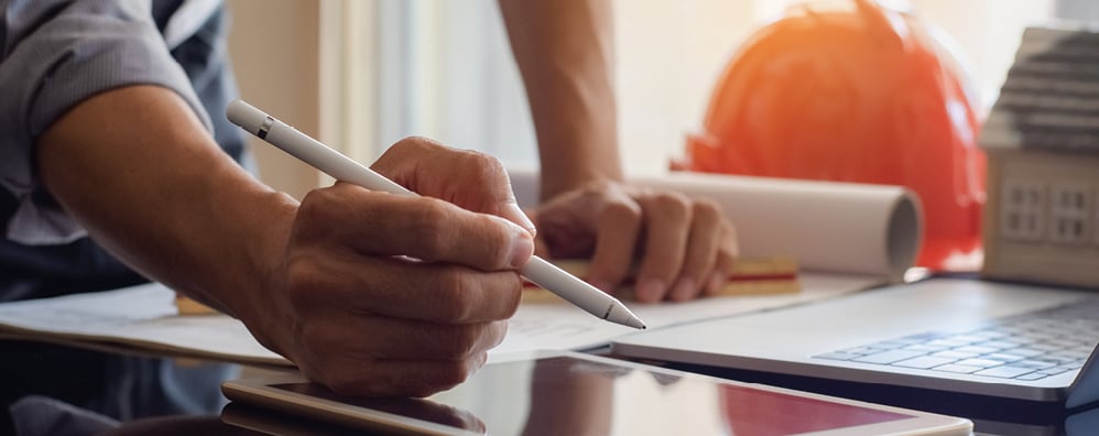 Person holding a pen over documents on a table, with a tablet, laptop, and orange hard hat in the background.