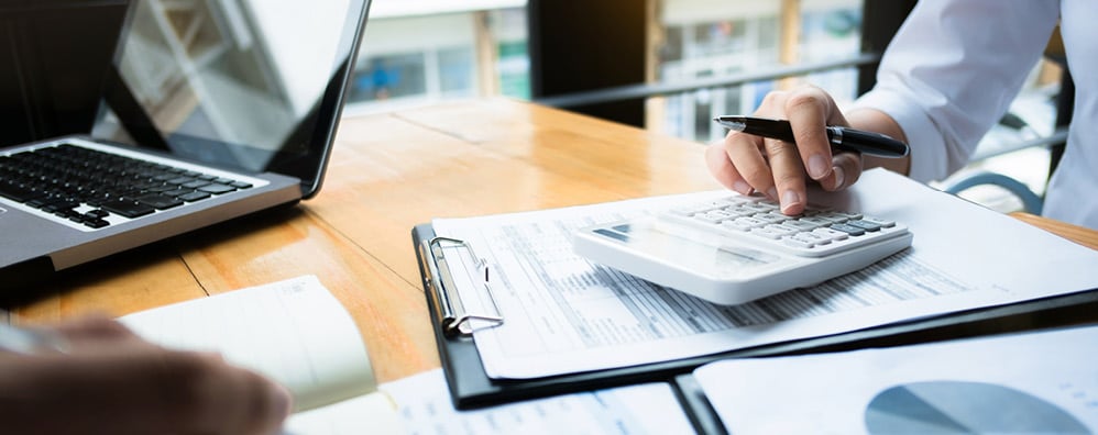 A person uses a calculator while holding a pen, with paperwork and a laptop on the desk.