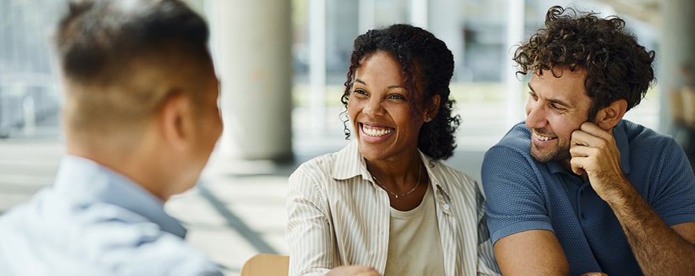 A woman smiles while shaking hands with a man across the table. Another man sits beside her, also smiling. The setting appears to be a bright office.
