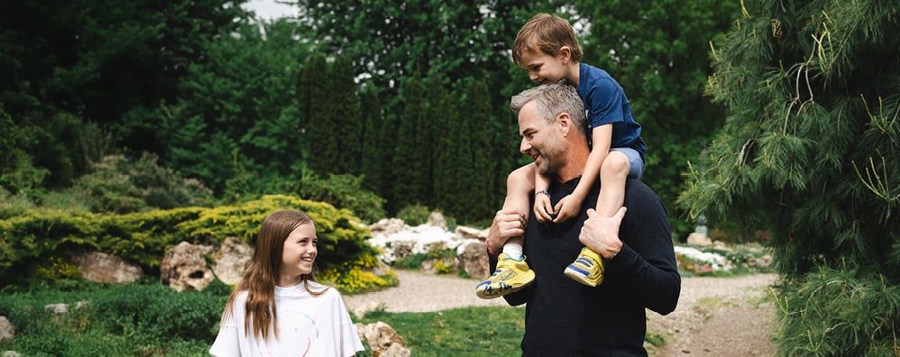 A dad with son on his shoulders while looking at his daughter carrying a football; the family is on a hike and green trees and rocks can be spotted behind them.