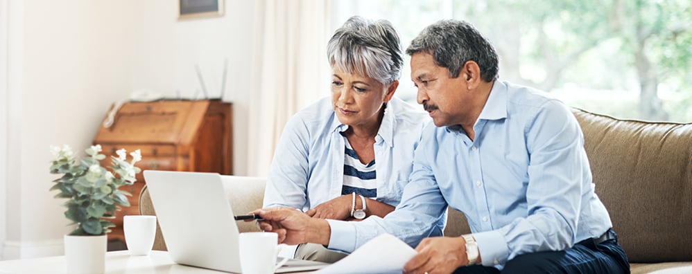 Elder couple sitting on a sofa in their living room as they look at their laptop; the male has papers in his hand while pointing at the laptop with a pen in his other hand.