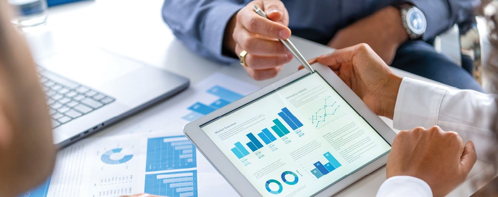 A business meeting with hands pointing at graphs on a tablet, showing blue bar and pie charts. 