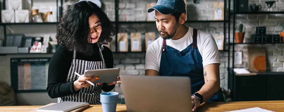 Two people in aprons, one holding a tablet and smiling while the other is typing on a laptop while looking over to the table. The two collaborate at a cozy cafe-style workbench.