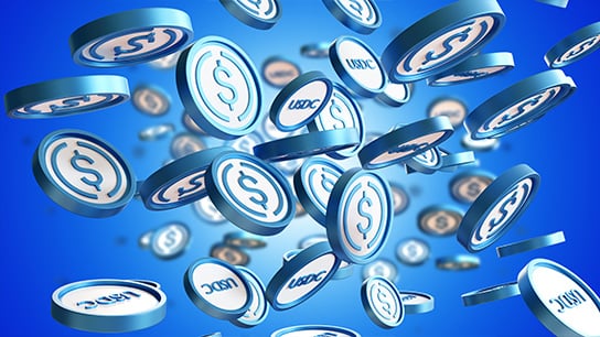 Numerous white coins with blue outline falling in front of a blue background. Some coins have a dollar sign while others have "USDC" 