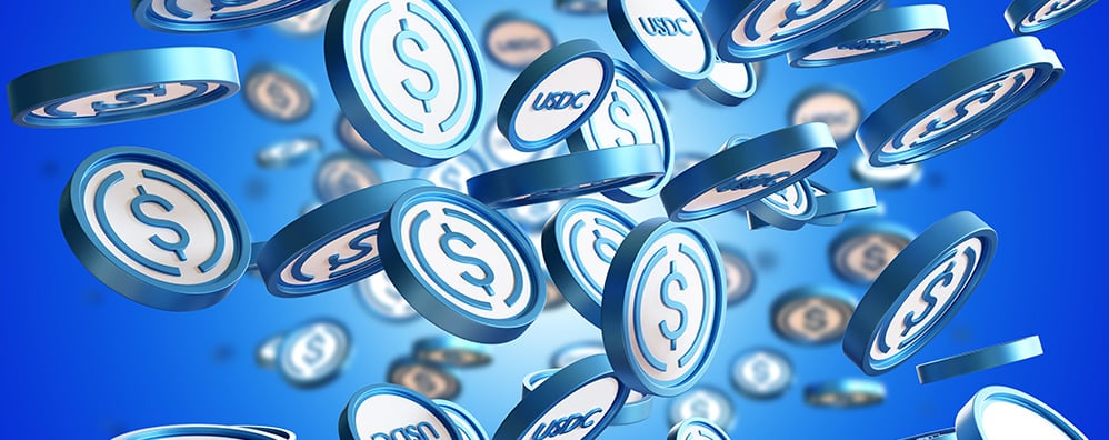Numerous white coins with blue outline falling in front of a blue background. Some coins have a dollar sign while others have "USDC" 