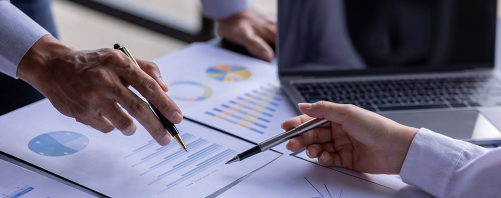 Two people going over financial charts on paper next to a laptop. Both are holding pens, and one is pointing with a pen, suggesting analysis or decision-making in an office setting.