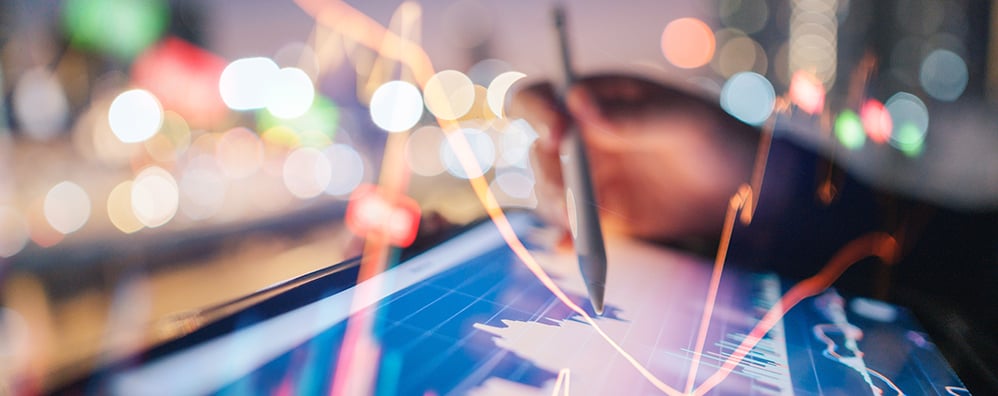 A hand holding a pen hovers over a digital tablet displaying financial graphs with glowing lines. The blurred background conveys a dynamic, analytical atmosphere.