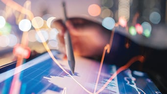 A hand holding a pen hovers over a digital tablet displaying financial graphs with glowing lines. The blurred background conveys a dynamic, analytical atmosphere.