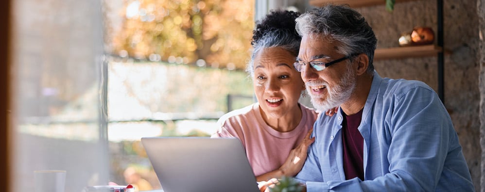 Elderly couple smiling and looking at a laptop in a bright room. The woman touches the man's shoulder, suggesting a warm and joyful moment.