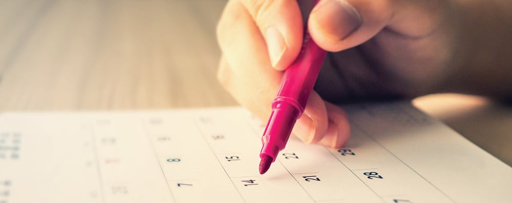 Hand holding a pink marker above a calendar, poised to mark a date.