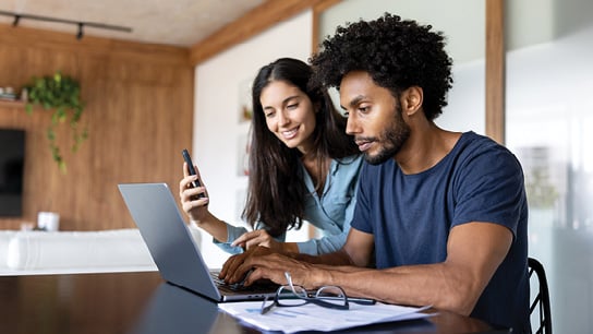 A woman and man collaborate together while looking at a laptop with surrounding documents on the table.. The man is sitting in front of the laptop while the woman is off to the side holding a phone up. 