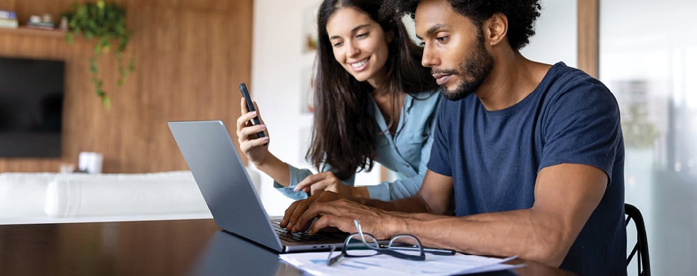 A woman and man collaborate together while looking at a laptop with surrounding documents on the table.. The man is sitting in front of the laptop while the woman is off to the side holding a phone up. 