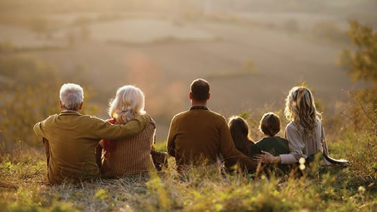 A multi-generational family of six sit together in a row on a grassy hill, facing the sunset. The grandparents sit on the left side while a couple with their two kids sit next to them.