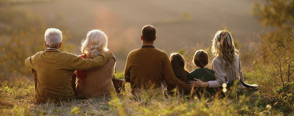 A multi-generational family of six sit together in a row on a grassy hill, facing the sunset. The grandparents sit on the left side while a couple with their two kids sit next to them.