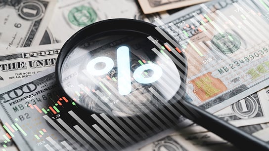 Magnifying glass with percent symbol in the center, while on top of cash bills. Transparent graphs overlay the image.