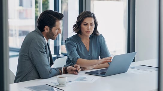 Woman and man in an office environment, sitting at a desk while looking at a laptop.