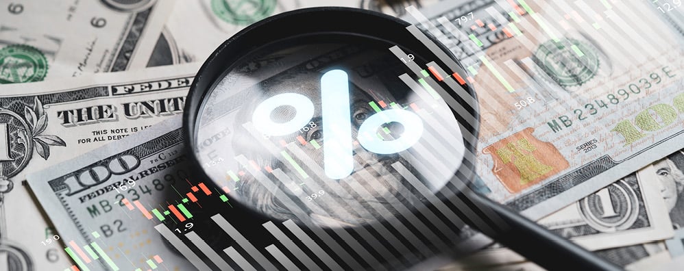 Magnifying glass with percent symbol in the center, while on top of cash bills. Transparent graphs overlay the image.