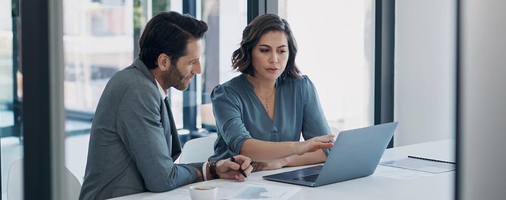 Woman and man in an office environment, sitting at a desk while looking at a laptop.