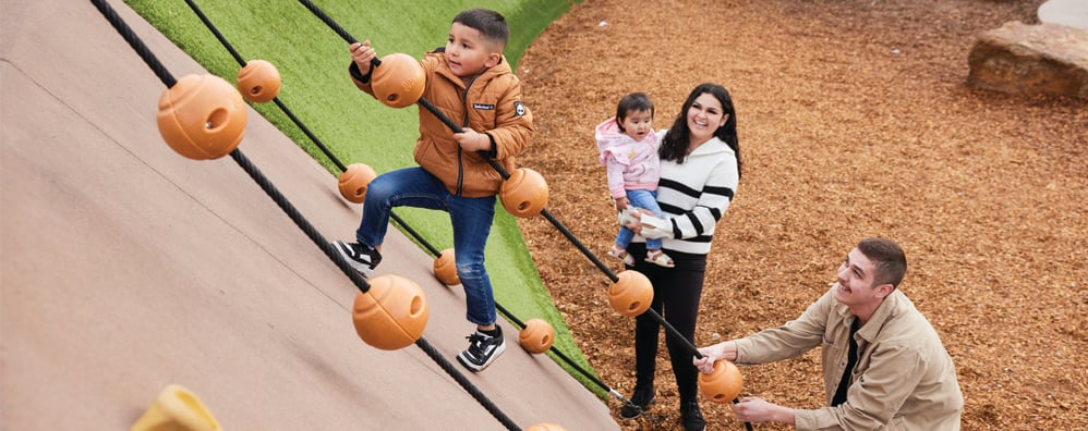 A child climbs a playground wall using ropes with orange grips, while a smiling woman holds a toddler and a man offers support, under a cheerful atmosphere.