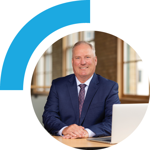 Middle-aged man smiling in a business suit while sitting at desk with laptop open.