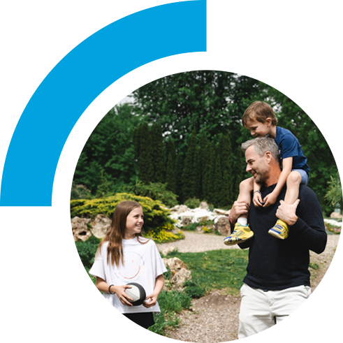 A dad has his son on his shoulders while smiling at his daughter. The family is on a hike with woods and rocks in the background.